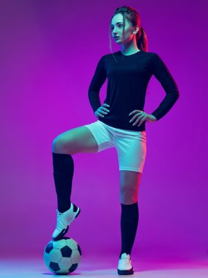 Portrait of female professional football player posing in uniform with ball isolated over purple background in neon